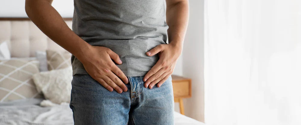 Man i vardagskläder med händerna på nedre delen av magen, stående vid en säng – fokus på bäckenområdet