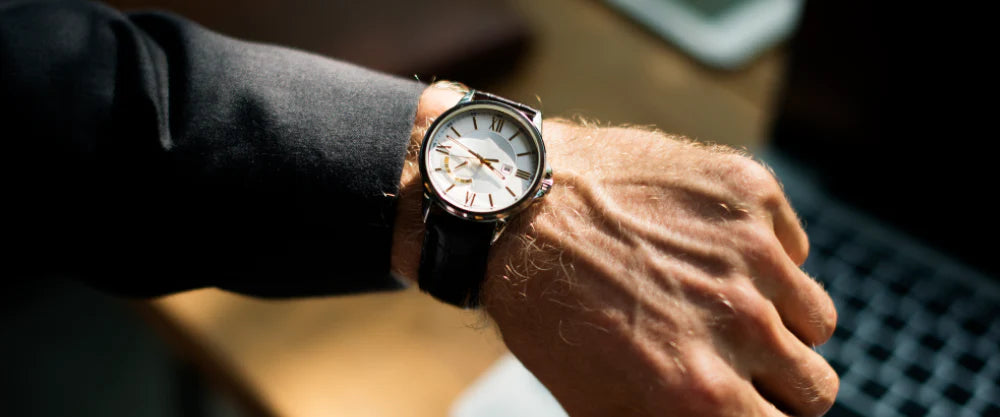 Man i kostym som tittar på sin armbandsklocka ovanför en bärbar dator – symbol för tid och kontroll