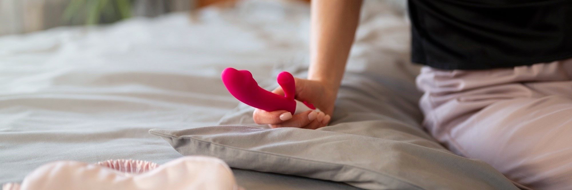En person sitter på en säng och håller i en rosa rabbit-vibrator med vit bas. Sängen har gråa sängkläder och i förgrunden syns ett plagg i ljusrosa färg.