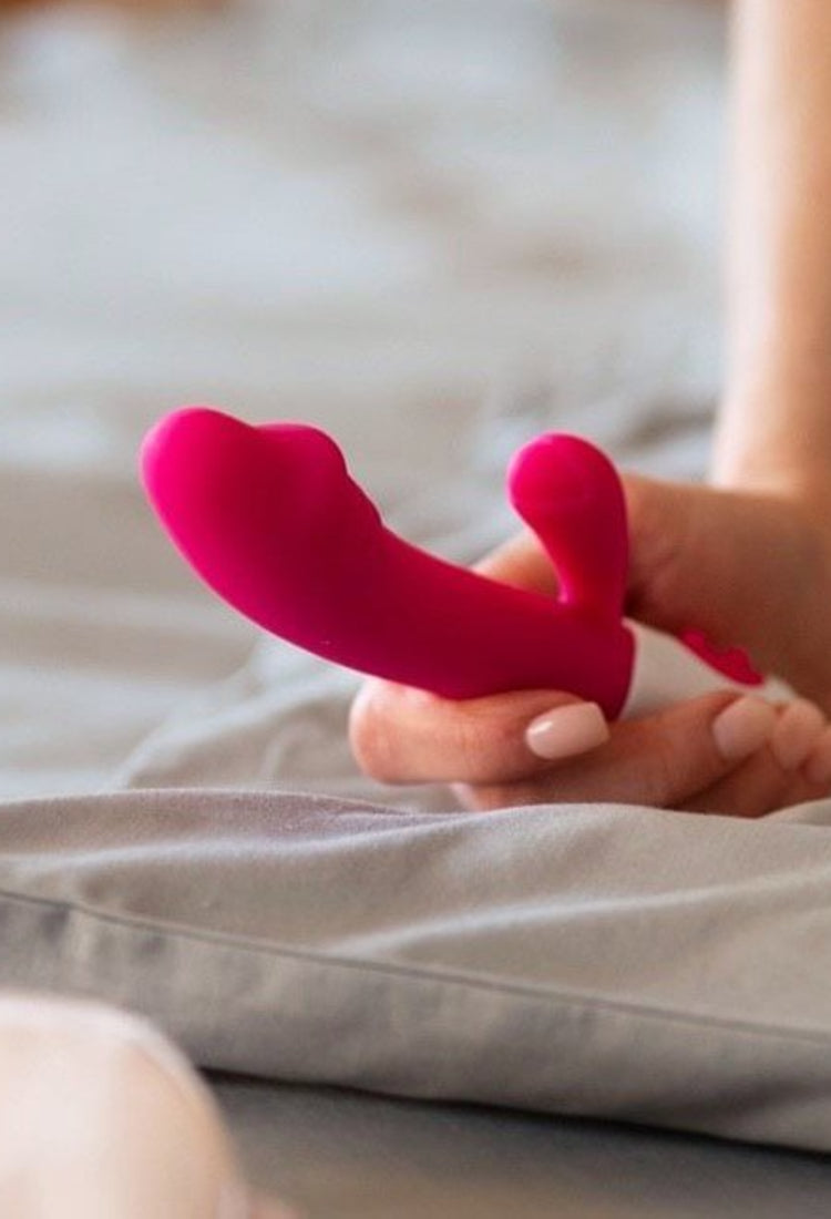 En person sitter på en säng och håller i en rosa rabbit-vibrator med vit bas. Sängen har gråa sängkläder och i förgrunden syns ett plagg i ljusrosa färg